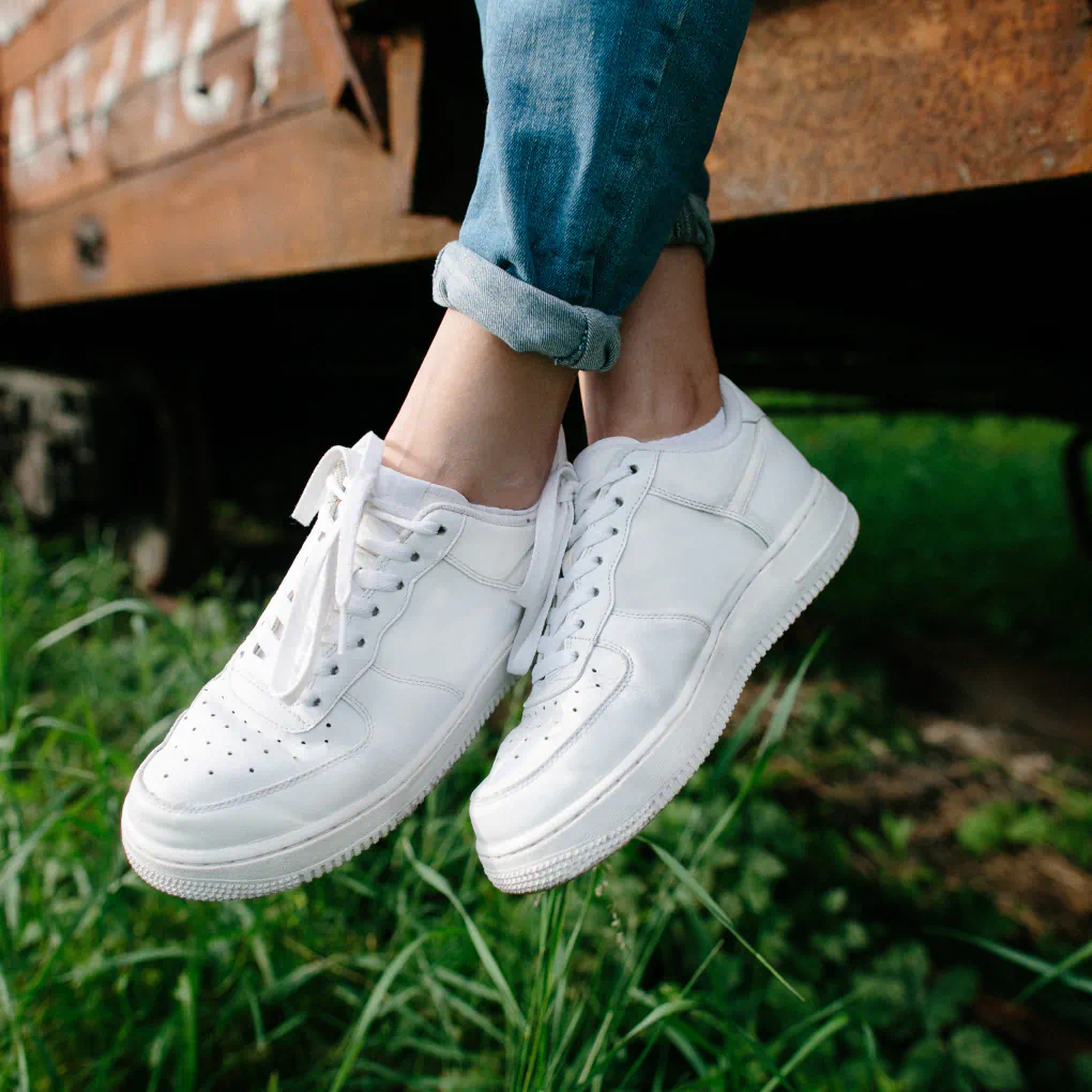 A person sits on a bench as their feet dangle toward a green lawn. They wear all-white sneakers.