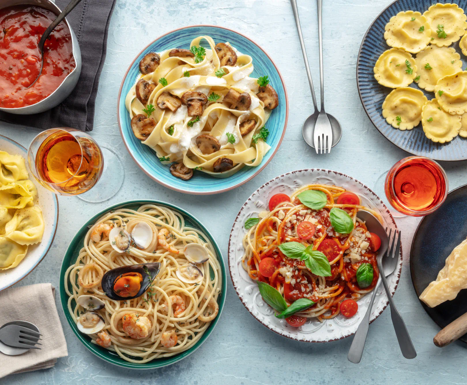 A spread of Italian food sits on a baby blue tablecloth. Shrimp pasta, spaghetti, pappardelle, ravioli, and rosé are all visible.