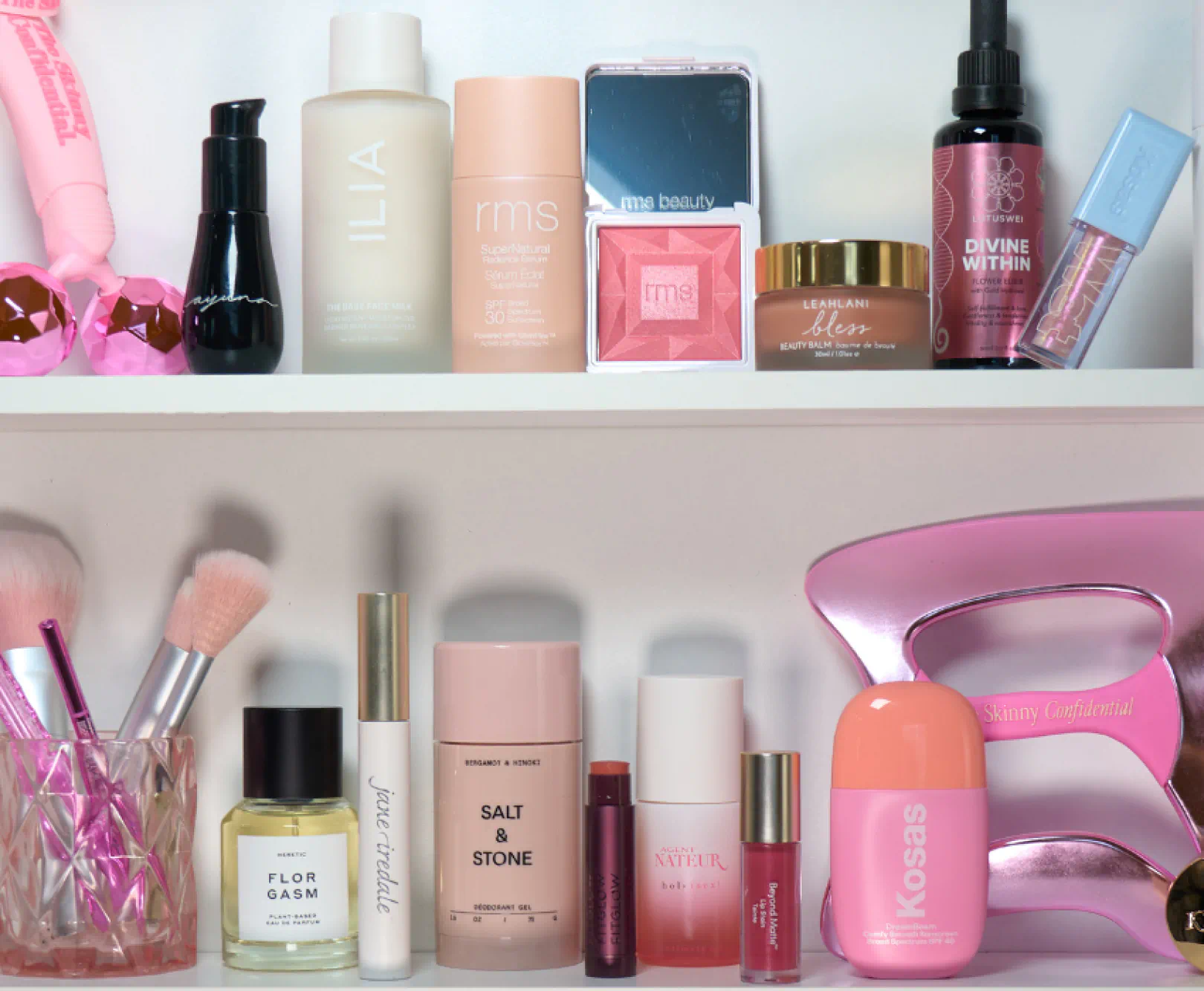 A collection of makeup supplies in pink and black bottles sit on two tiers of white shelves.