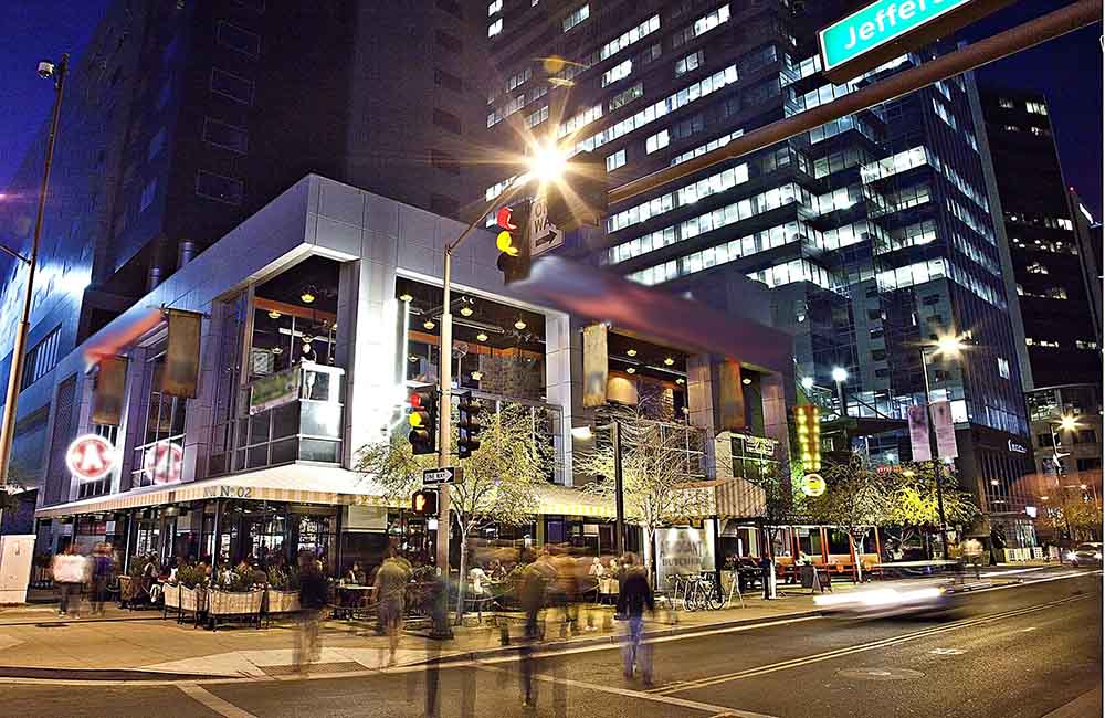Long exposure shot of an intersection at CityScape. The blurred image of people crossing in front of fancy store front with luxury spaces above and a large skyscraper in the background.