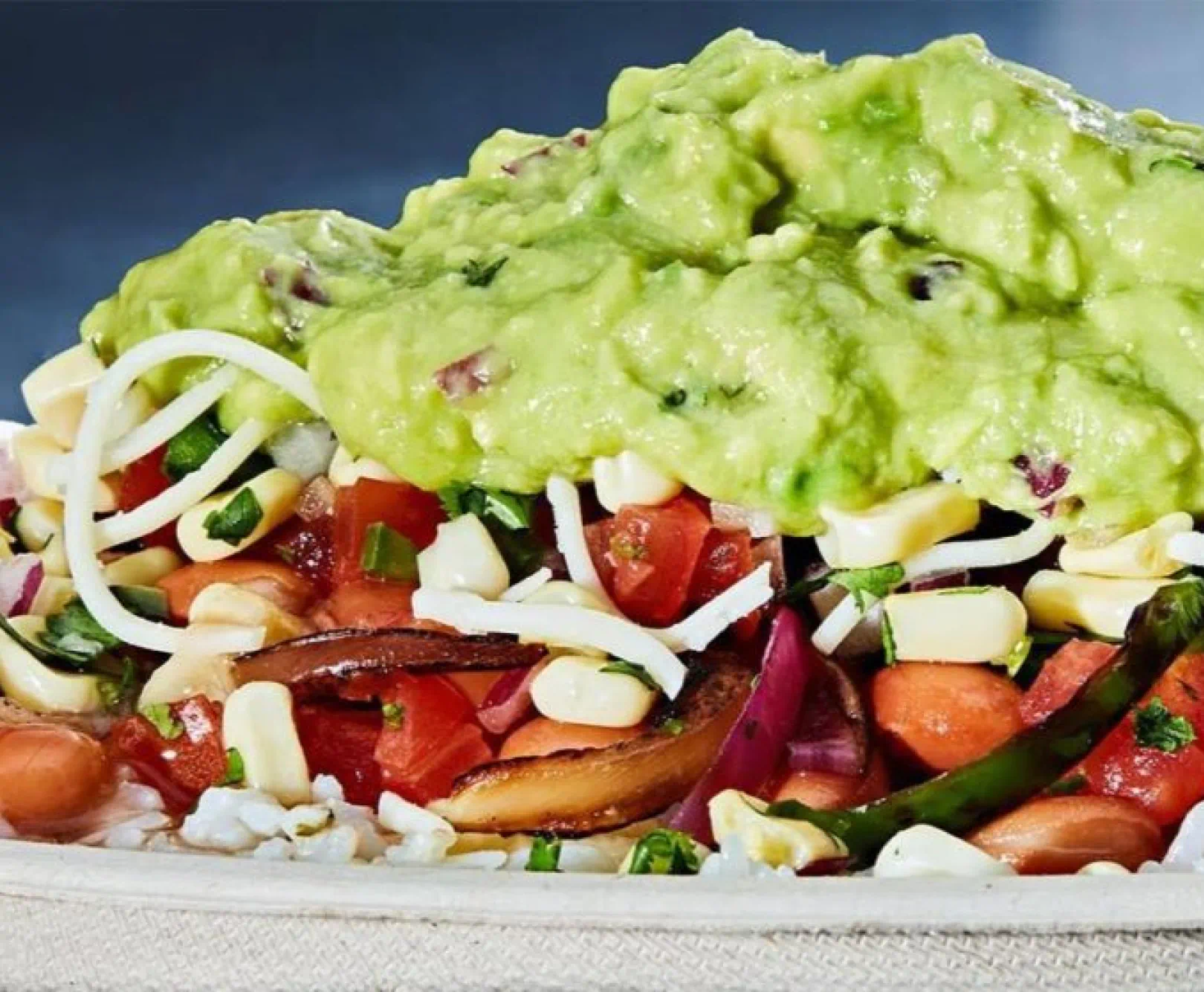 Close-up of a fresh food dish topped with guacamole, shredded cheese, diced tomatoes, corn, and sliced mushrooms.
