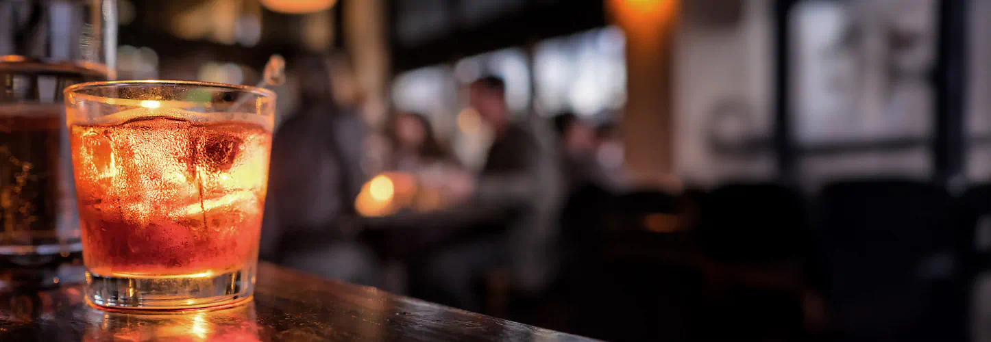A lone old fashioned cocktail sits on the bar, blurred people in a restaurant in the background.