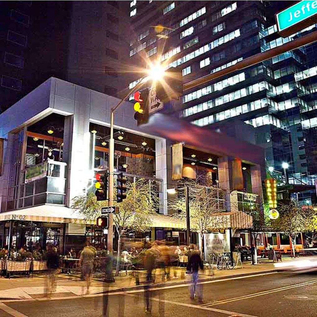 Long exposure shot of an intersection at CityScape. The blurred image of people crossing in front of fancy store front with luxury spaces above and a large skyscraper in the background.