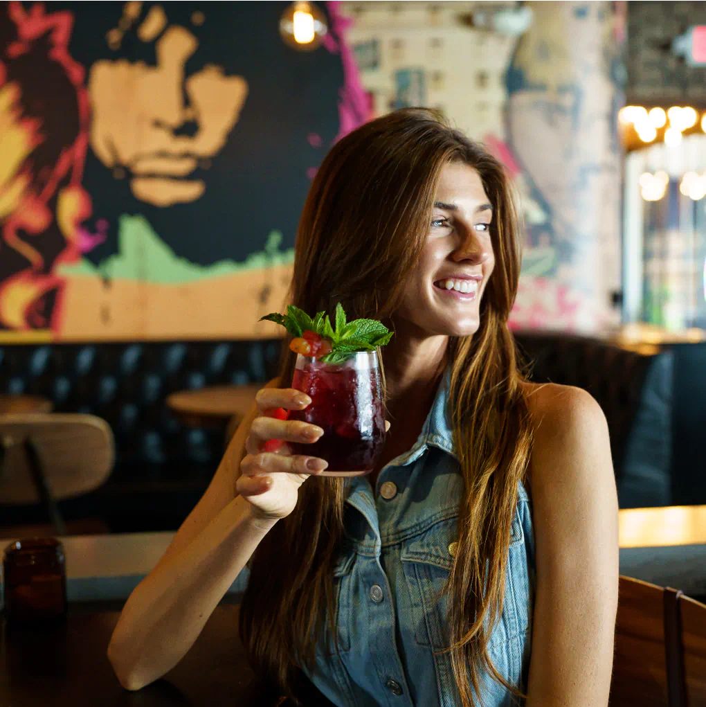A smiling woman sits in a restaurant holding a deep red colored cocktail garnished with mint.