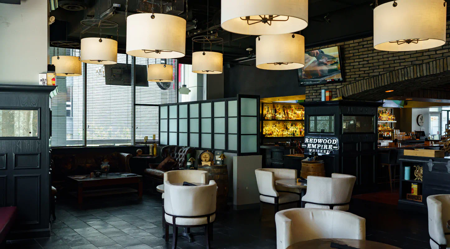Interior of a lobby with large lights suspended from the ceiling. Small tables are in the foreground and behind a divider in the background there is an entrance to a bar.