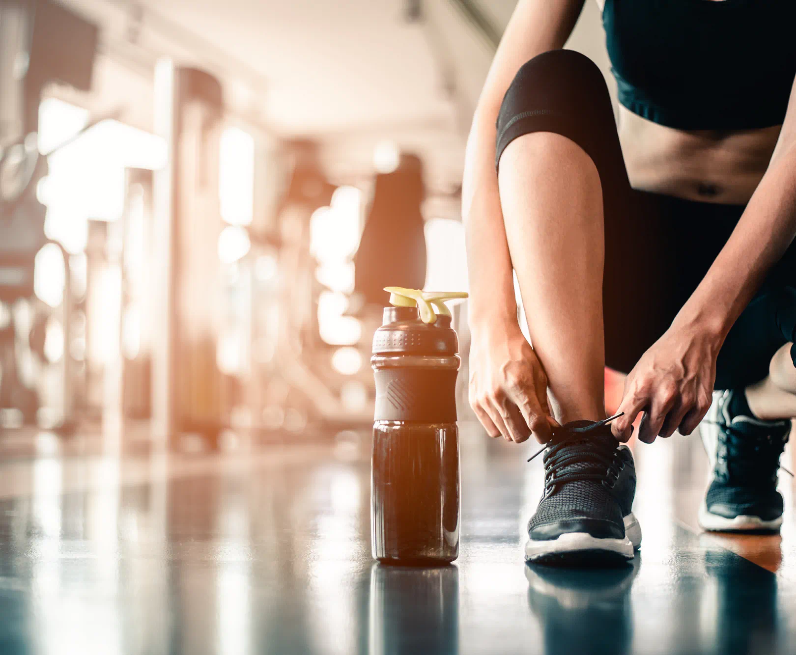 a woman in athletic wear adjusts her running shoes in a gym with a black water bottle sitting beside her