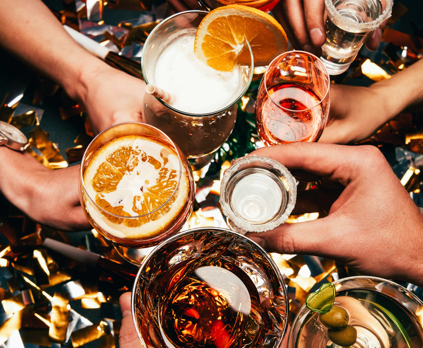 Hands holding various drinks over a table with golden confetti.