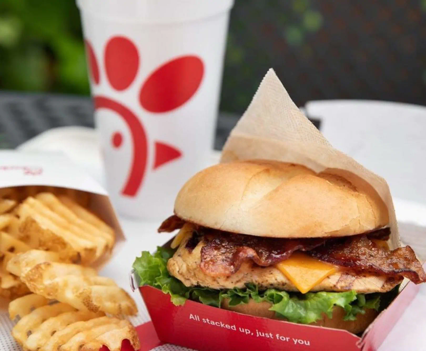 a chick-fil-a chicken sandwhich with bacon, lettuce and cheese. beside it sits a container of waffle fries and a soft drink cup bearing the chick-fil-a chicken logo. the sandwich container text reads "all stacked up, just for you"