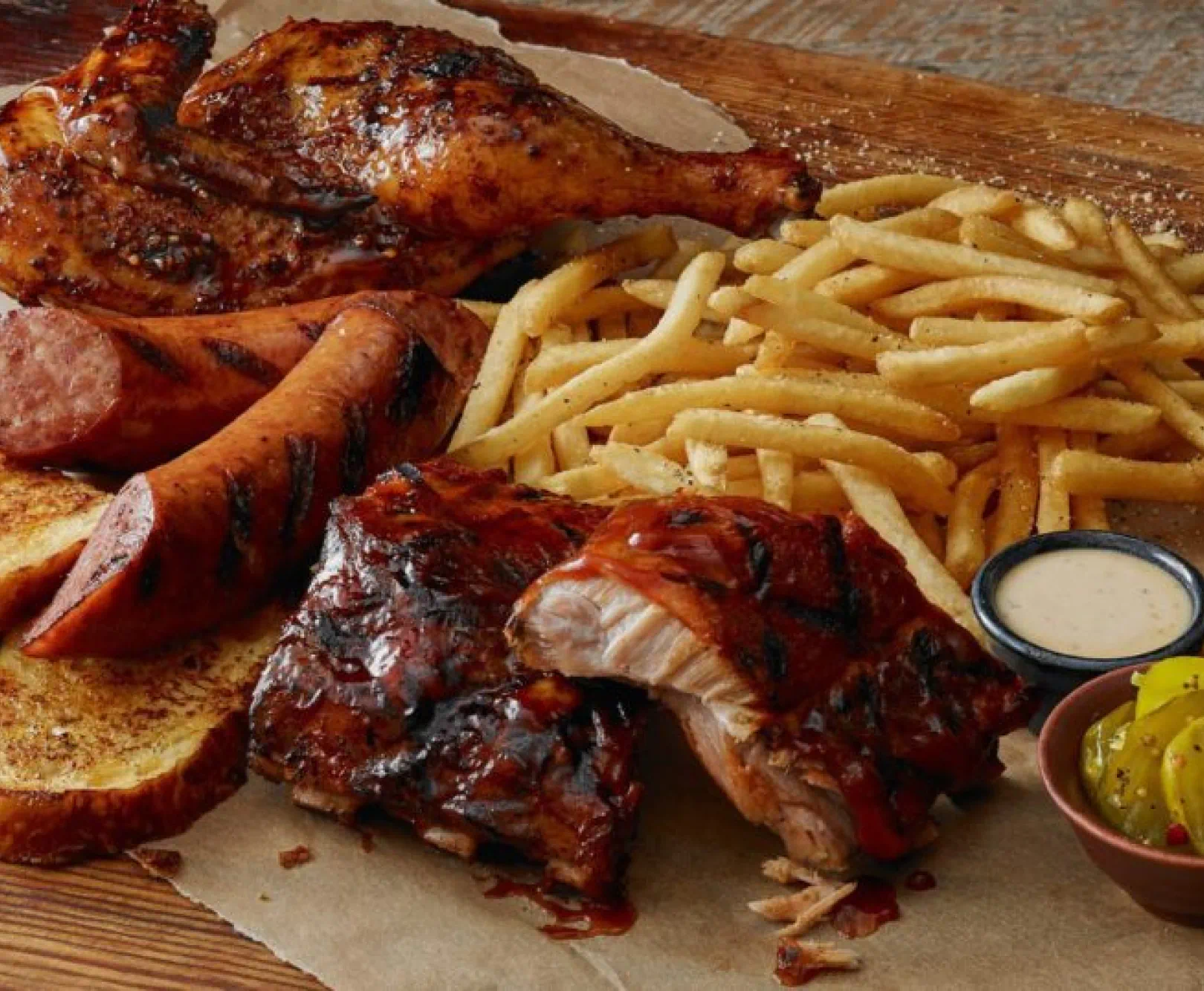 Grilled chicken, sausages, barbecued ribs, French fries, grilled toast, a dipping sauce, and sliced pickles on a wooden platter.
