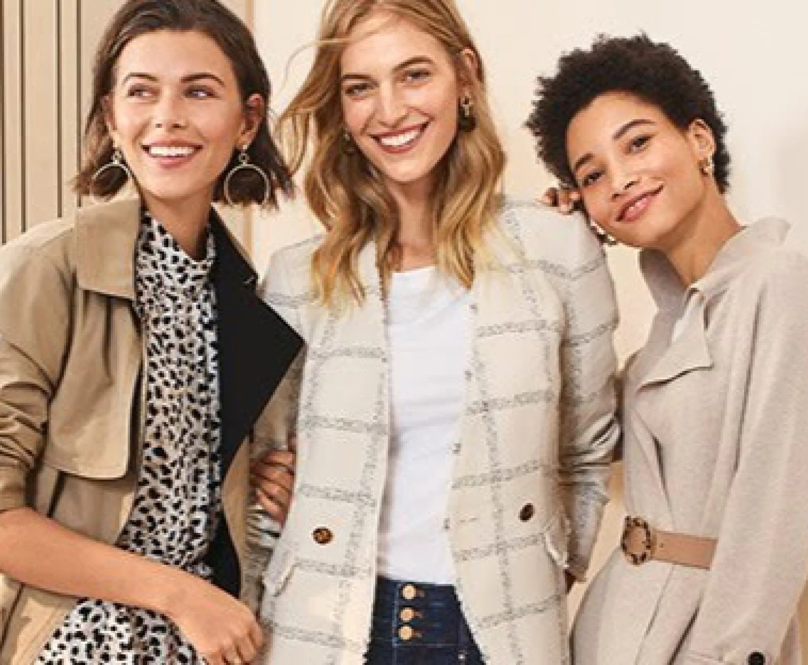 Three women smile as they show off stylish coats in tan and blue.