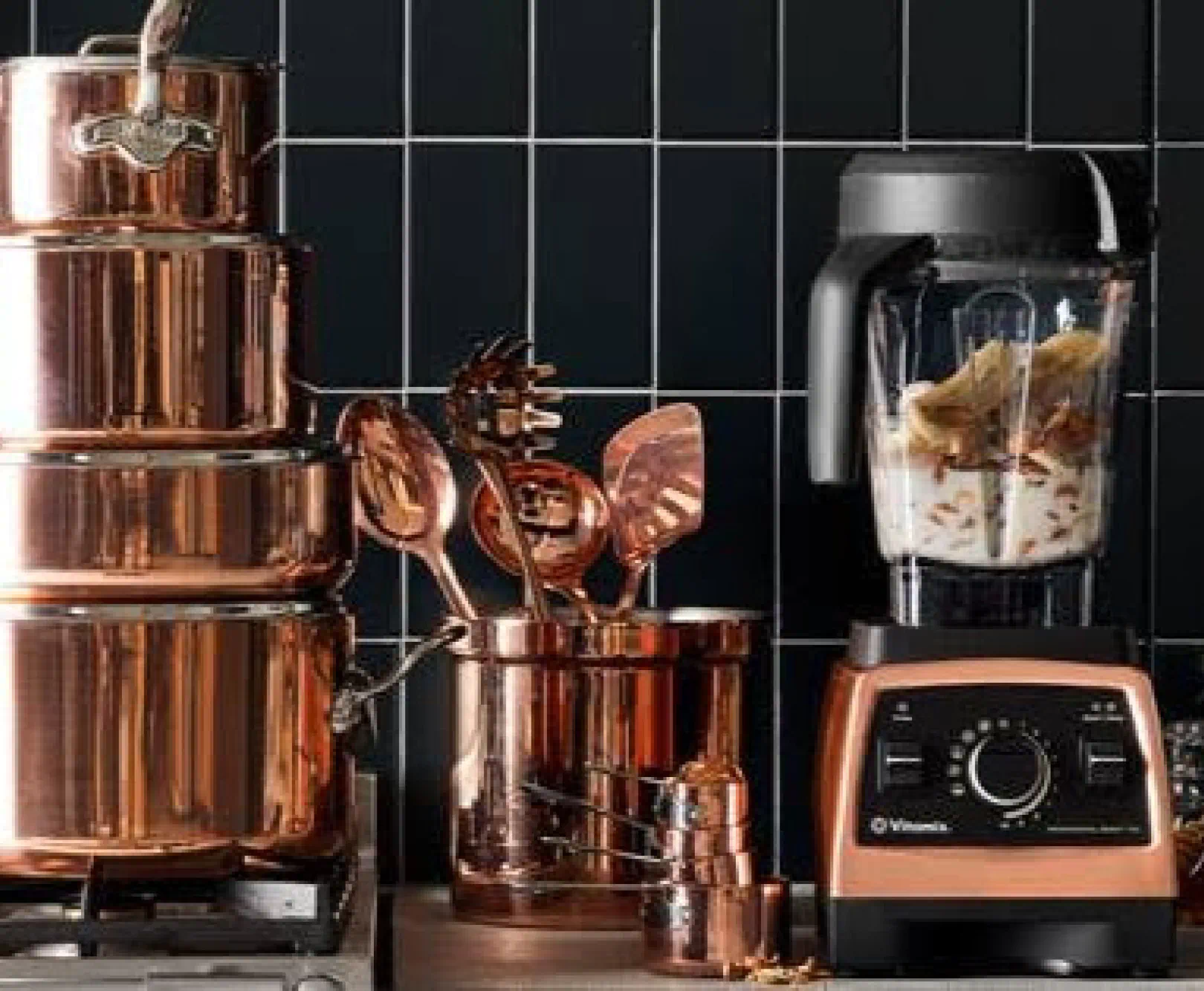 A group of copper kitchenware sits in front of a black tiled wall.