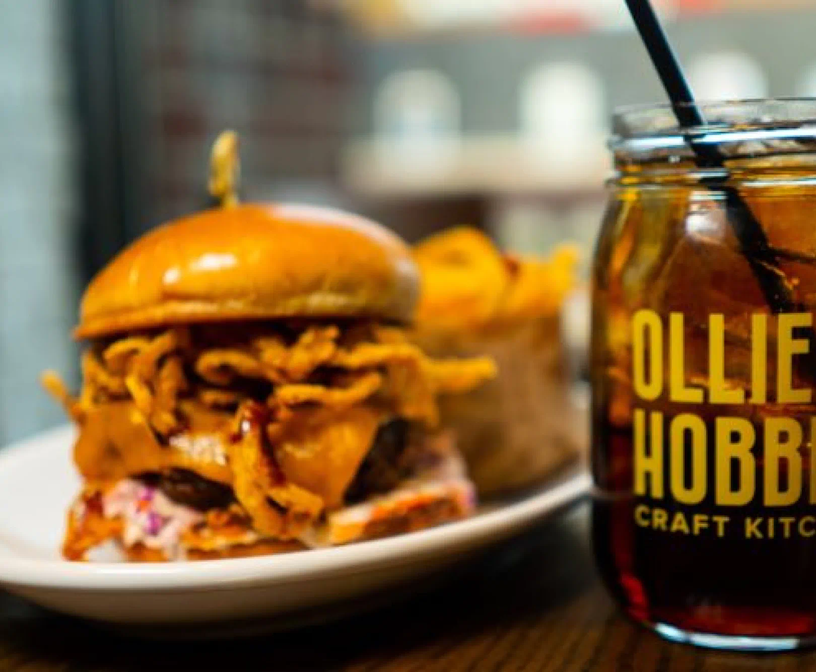 Gourmet burger with fried onions and cheese, accompanied by fries and a drink in a labeled jar.