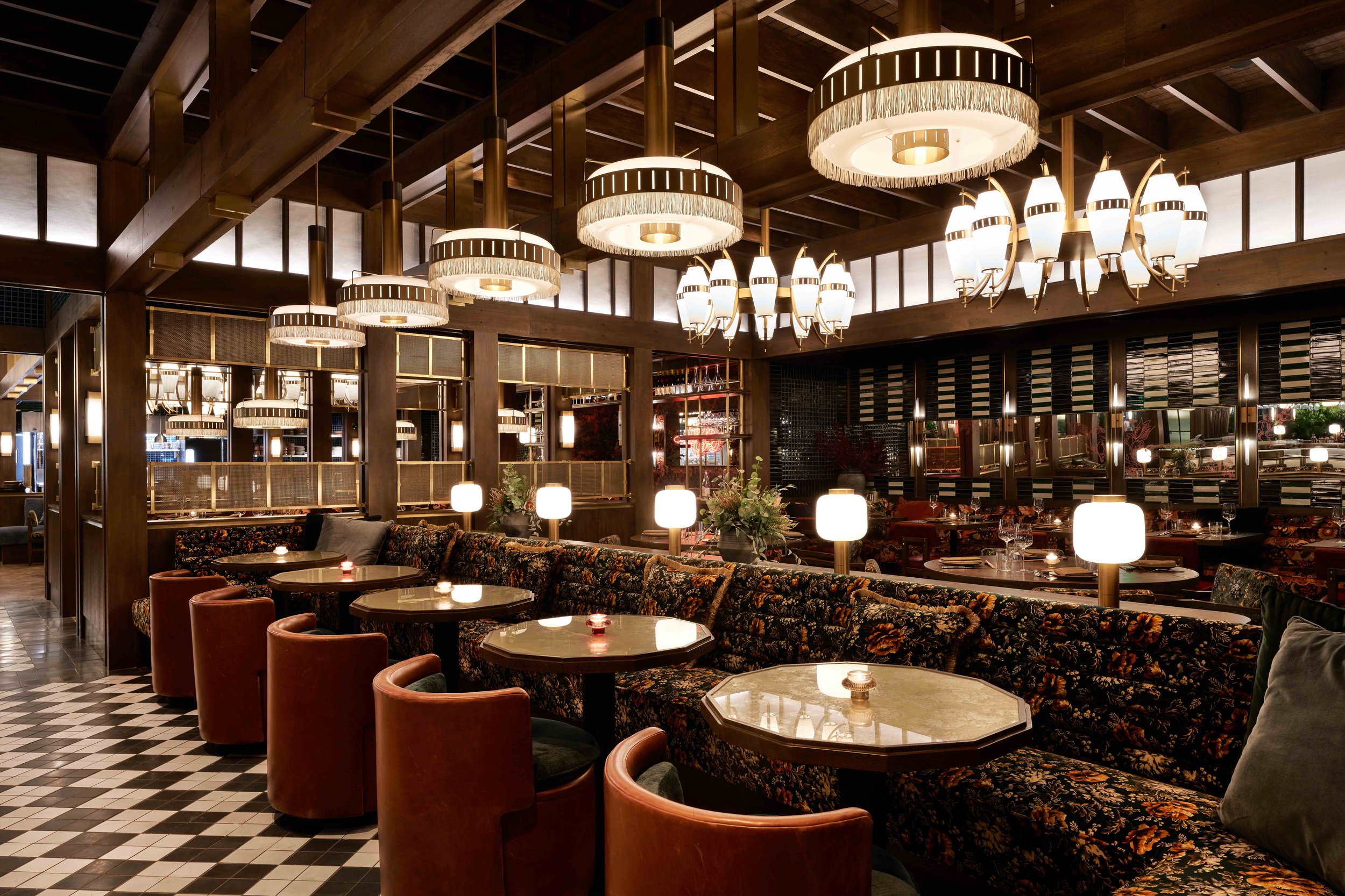 Interior of a stylish bar with large light fixtures hanging from wooden beams on the ceiling. A long both made of a floral print is lined with a row of small tables and orange chairs.