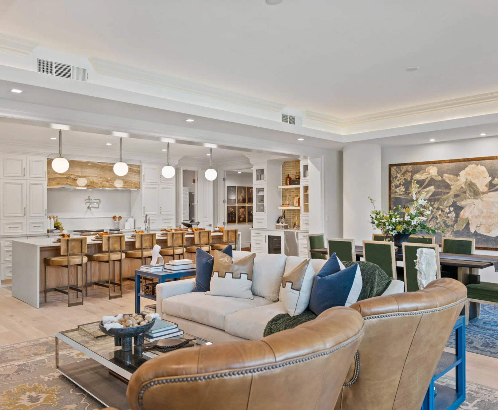 Open-concept living area with a beige sectional sofa, brown leather armchairs, a glass coffee table, and a kitchen island with white cabinetry and pendant lights in the background.