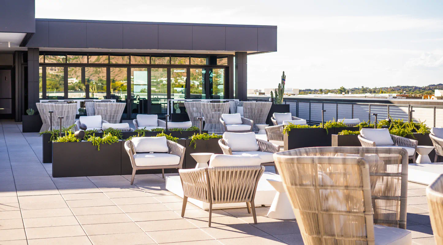 Outdoor rooftop lounge area with modern furniture, and a dark grey building with large glass windows in the background.