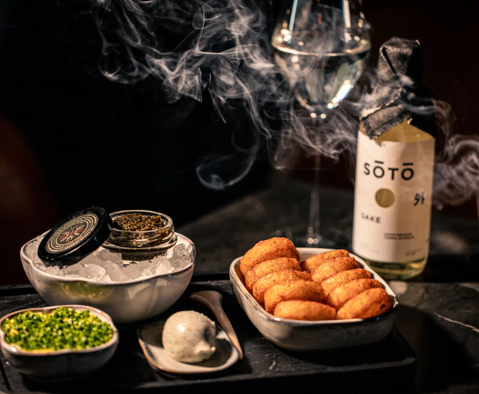 An elegant arrangement of appetizers, herbs, caviar, and a bottle of SOTO sake with a glass.