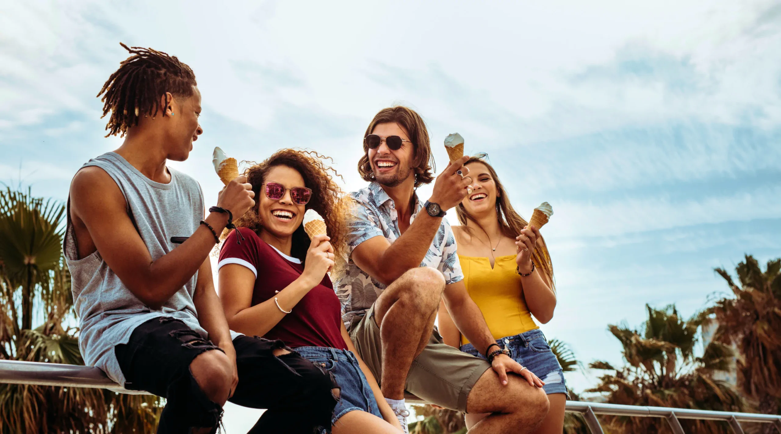 A group of young, smiling people are enjoying ice cream in cones. Palm trees sit in the background.